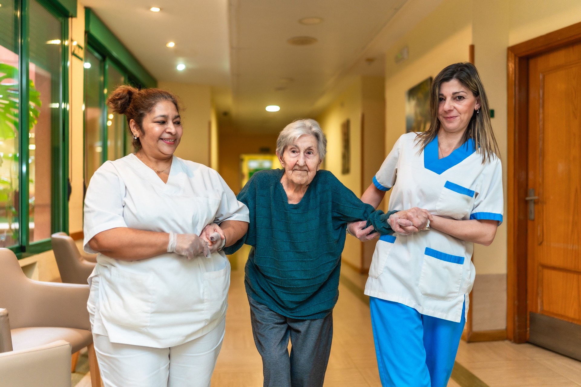 Nurses helping elderly woman walking in nursing home corridor